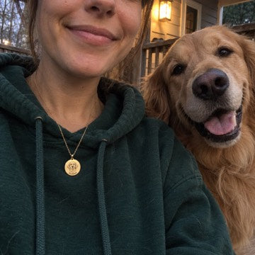 AI concept: A gold necklace engraved SUNNY is worn over a green hoodie beside the matching golden retriever on a porch.