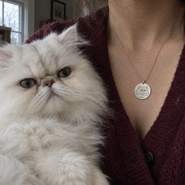 Illustration: White Persian Luna beside a worn silver portrait necklace against a burgundy cardigan.